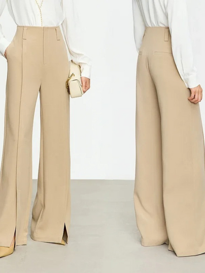 A woman showing the front and back of a pair of cream colored, tailored, pleated, high waisted pants with slits at the hem.