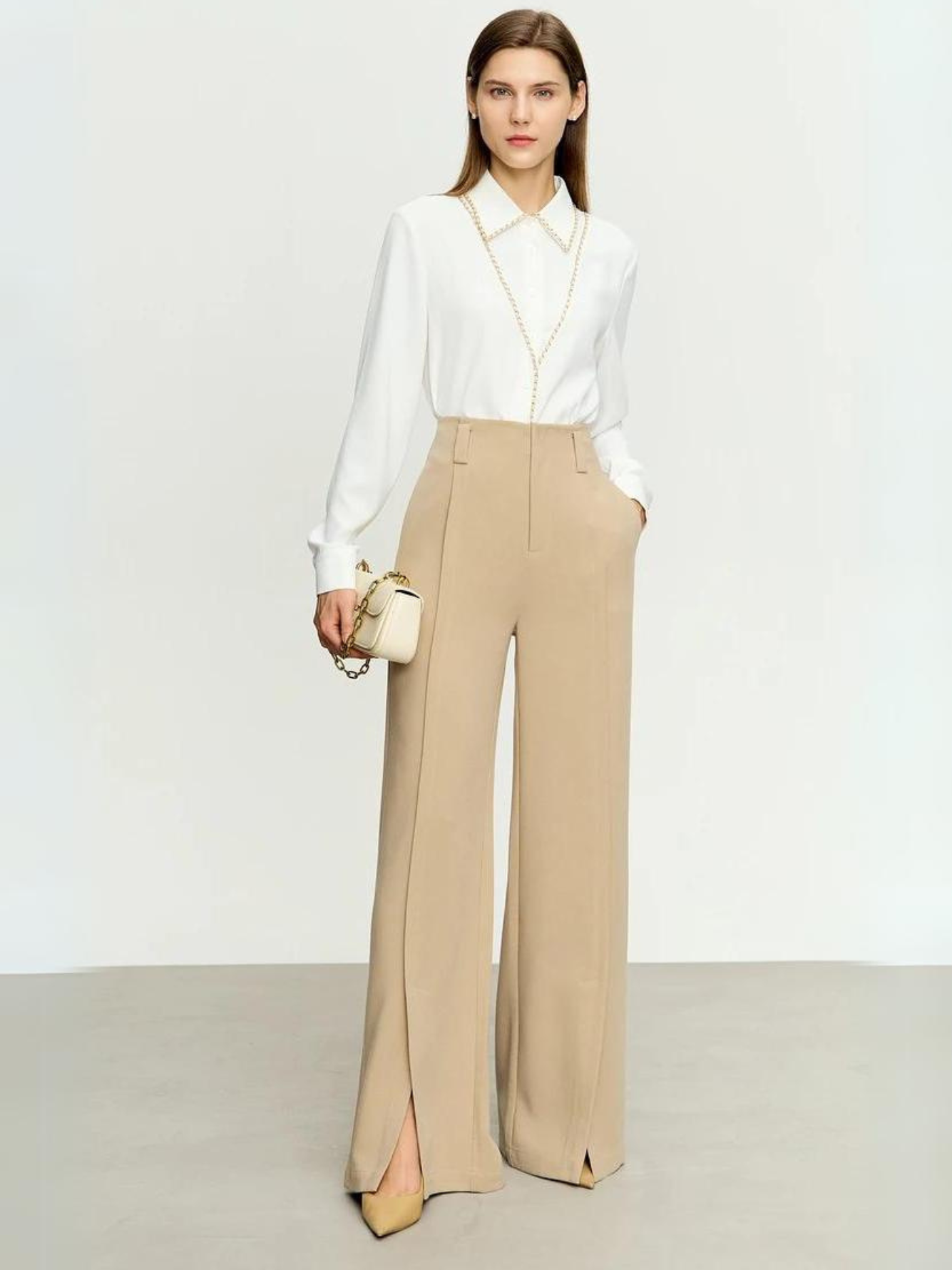 A woman wearing tailored, pleated, high waisted pants with slits at the hem with a simple white long-sleeved blouse and bag.