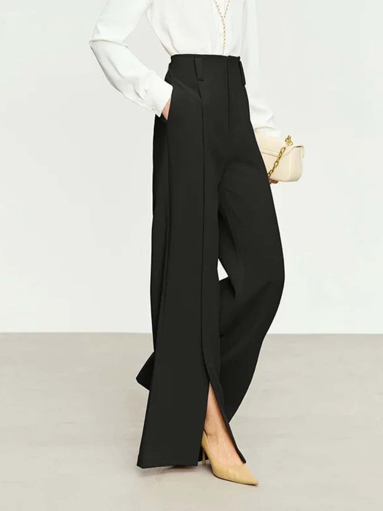 A woman wearing a pair of black, tailored, pleated, high waisted trousers with slits at the hem with a simple white long-sleeved blouse and bag.