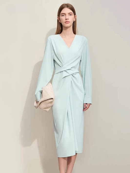 A woman wearing a light blue, mid-calf length, long sleeved, flowy dress with a v-neckline and a draping design on the bodice and a bow on the back to cinch the waist.