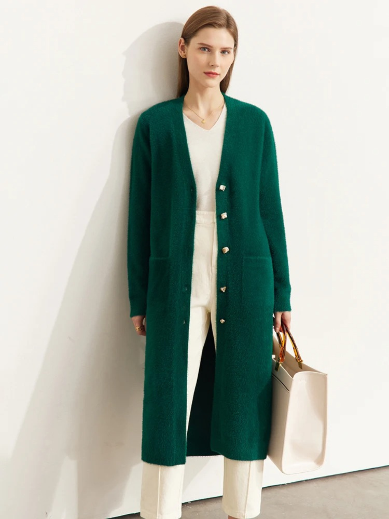 A brunette woman wearing a long, deep green cardigan over a white shirt and beige tailored pants.