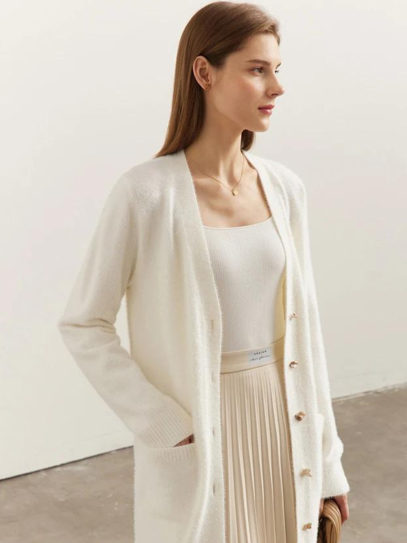 A brunette woman wearing a long, cream colored cardigan over a cream colored pleated skirt with her right hand inside the deep pocket of the cardigan.