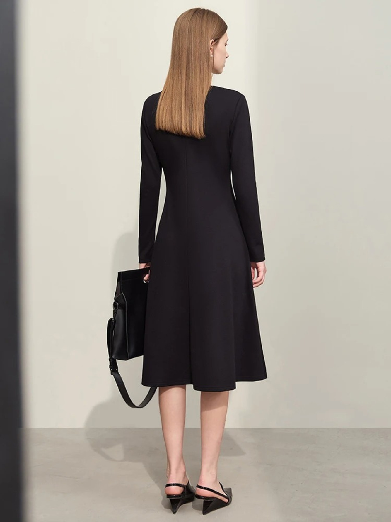 A woman showing the back of a black, long sleeve, below the knee dress with an a-line skirt paired with black slingback pumps and a black bag.