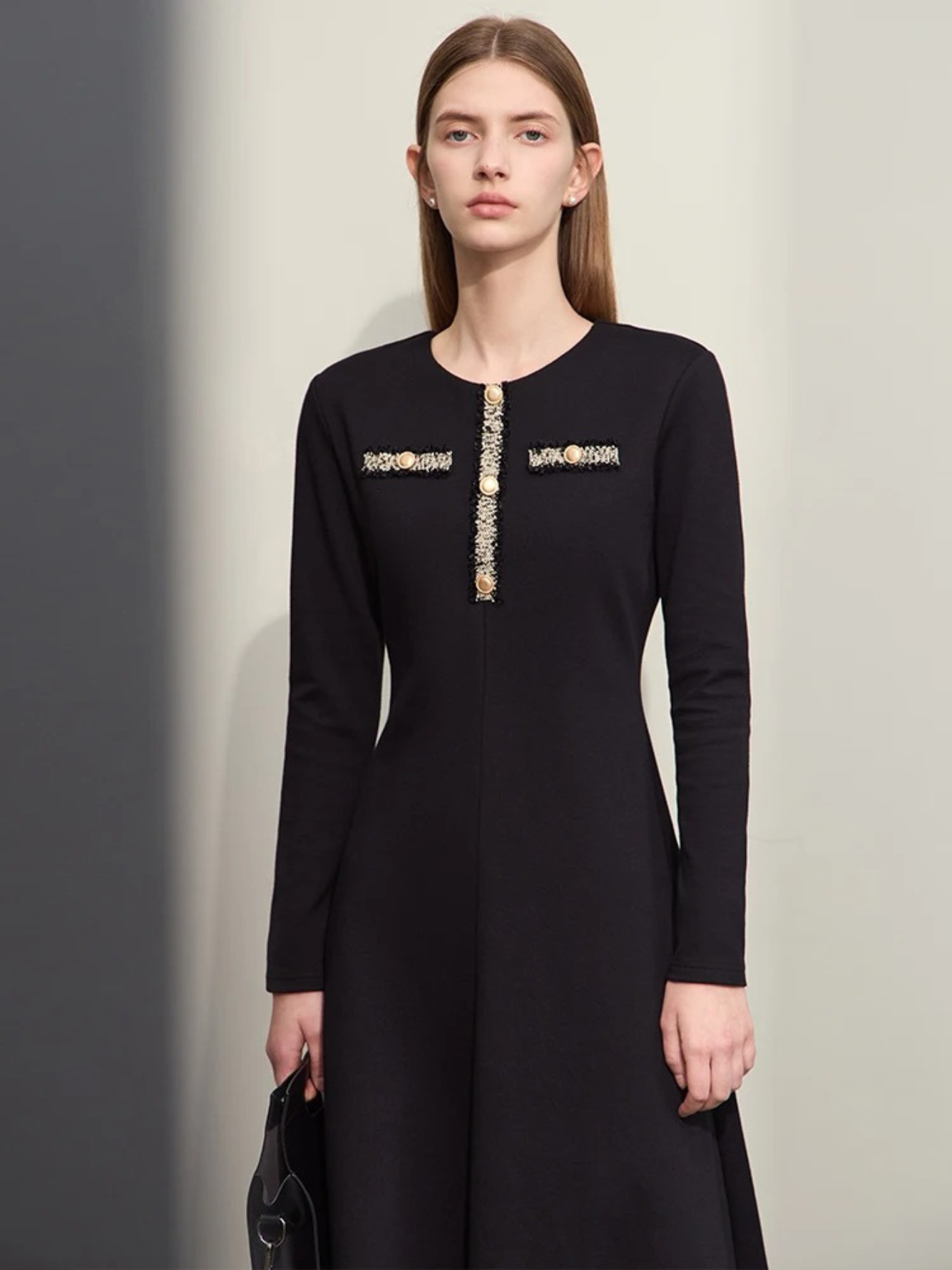 A woman wearing a black, long sleeve, below the knee dress with embroidery and button design on the chest and an a-line skirt paired with a black bag.