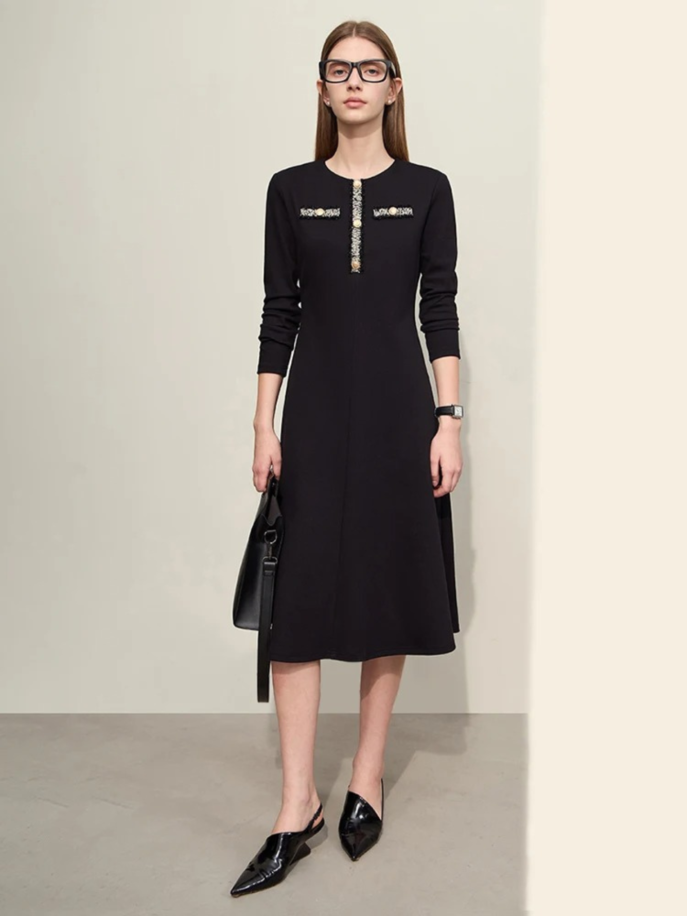 A woman wearing a black, long sleeve, knee-length dress with an a-line silhouette. The dress has a round neckline and has an embroidery and gold button design on the chest. She is wearing black slingback heels and a black bag.