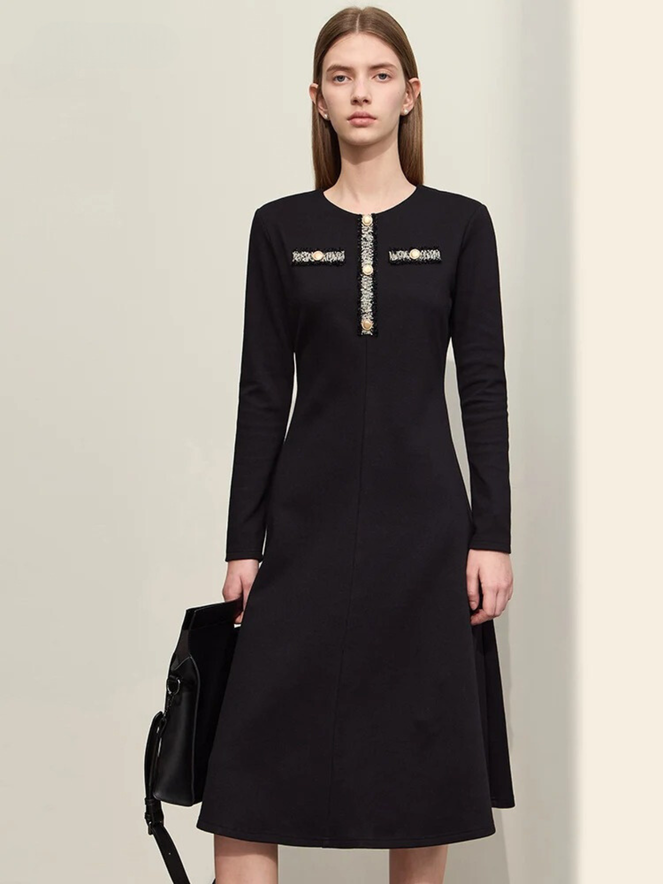 A woman wearing a black, long sleeve, knee-length dress with an a-line silhouette. The dress has a round neckline and has an embroidery and gold button design on the chest. She is also carrying a black bag.