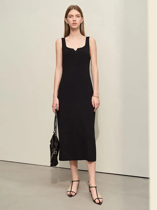 A woman wearing a minimalist, knee length, sleeveless dresses with thin straps and a mini cut out in the neckline in black paired with black strappy sandals and a bag.