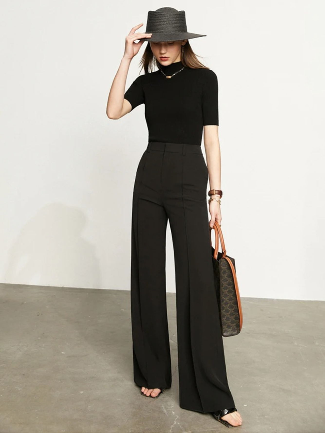 A woman wearing a pair of black, tailored, high waisted, floor length pants with a plain top, a handbag, and a stylish hat.