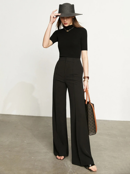 A woman wearing a pair of black, tailored, high waisted, floor length pants with a plain top, a handbag, and a stylish hat.