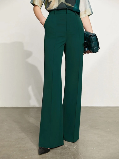 A woman wearing a pair of green, tailored, high waisted, floor length pants with slits at the hem with a printed blouse and handbag.