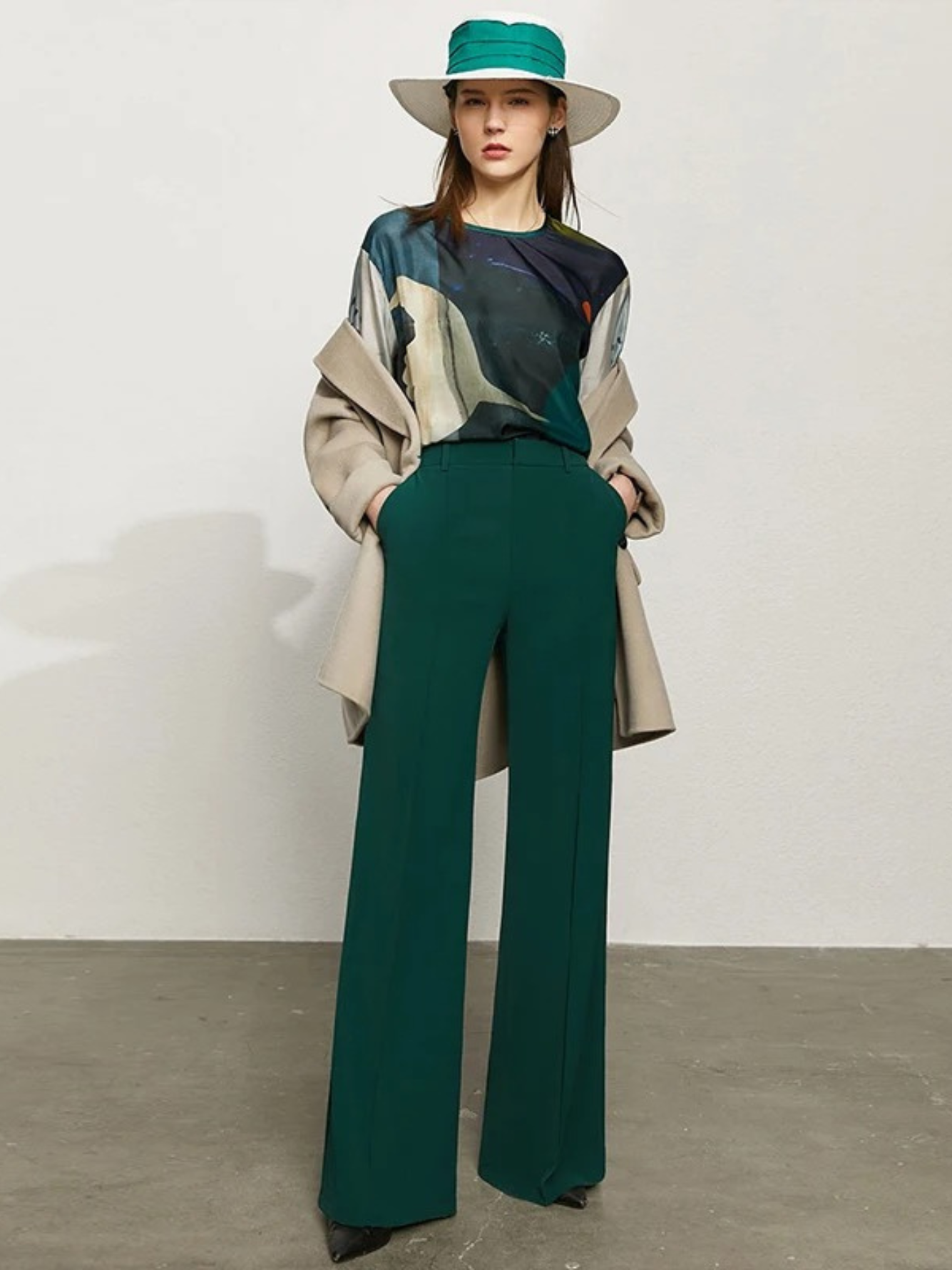 A woman wearing a pair of green, tailored, high waisted, floor length pants with slits at the hem with a printed blouse, a coat, and a hat.