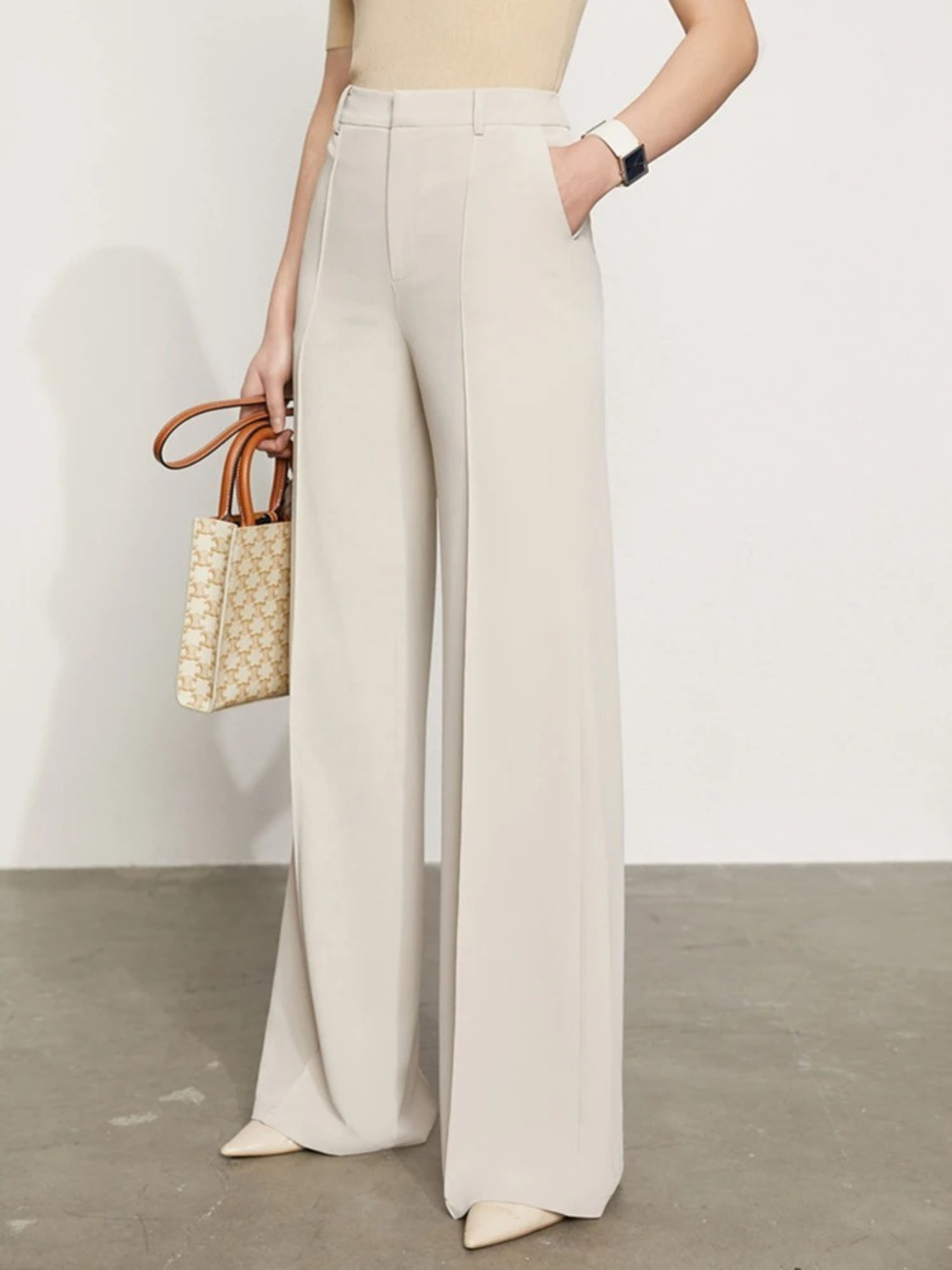 A woman wearing a pair of cream, tailored, high waisted, floor length pants with slits at the hem with a plain top and a stylish handbag.