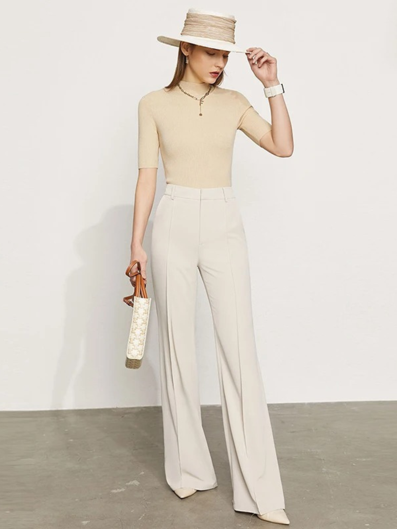 A woman wearing a pair of cream, tailored, high waisted, floor length pants with slits at the hem with a plain top, a hat, and a handbag.