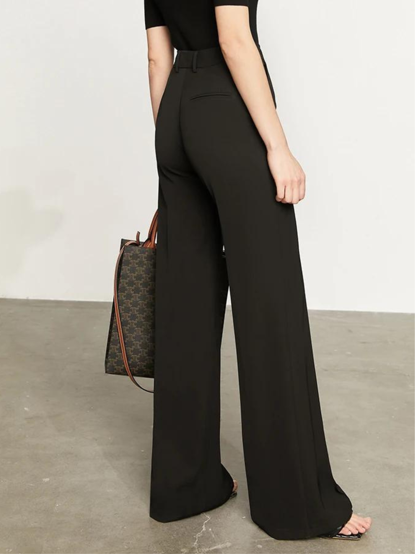 A woman showing the back of a pair of black, tailored, high waisted, floor length pants with a plain top and a handbag.