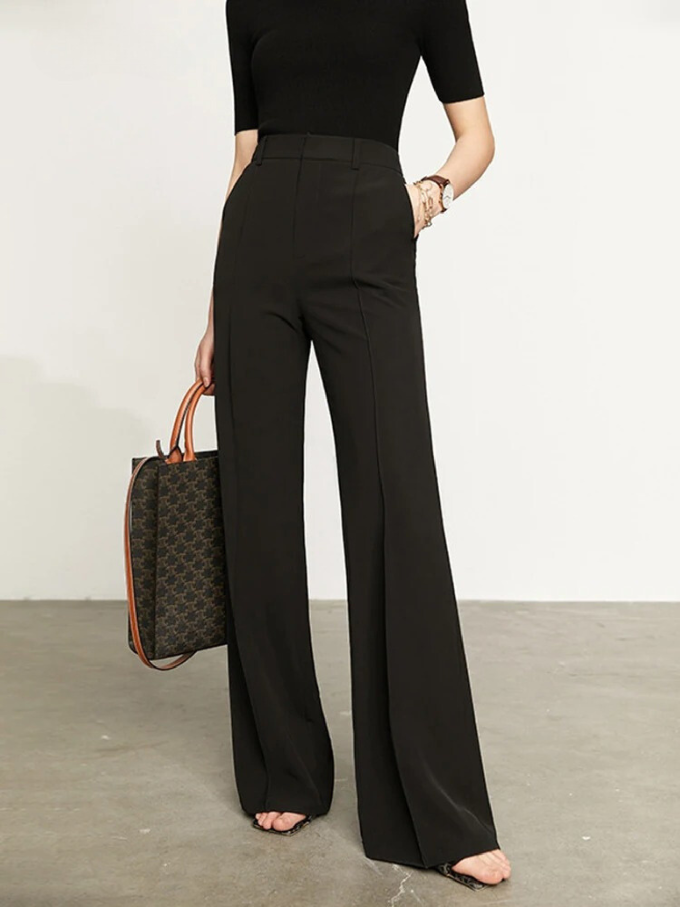 A woman wearing a pair of black, tailored, high waisted, floor length pants with a plain top and a handbag.