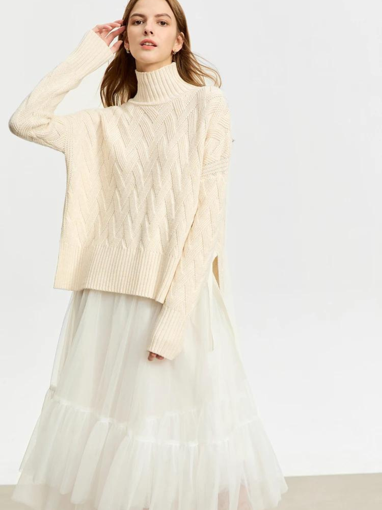 A woman wearing a cream colored knit turtleneck sweater with a tie design on both sides and a chevron knitting pattern and a white tulle skirt.